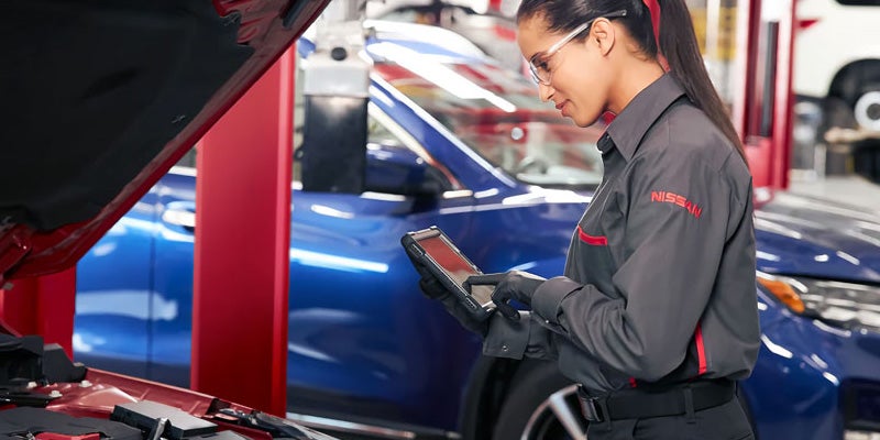 A woman in a gray mechanic's uniform examines a device while standing next to a Nissan car with its hood open in an auto service shop.