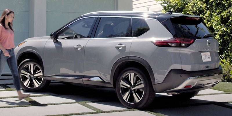 A woman walking past a gray used Nissan Rogue SUV parked in a driveway with a modern house in the background.