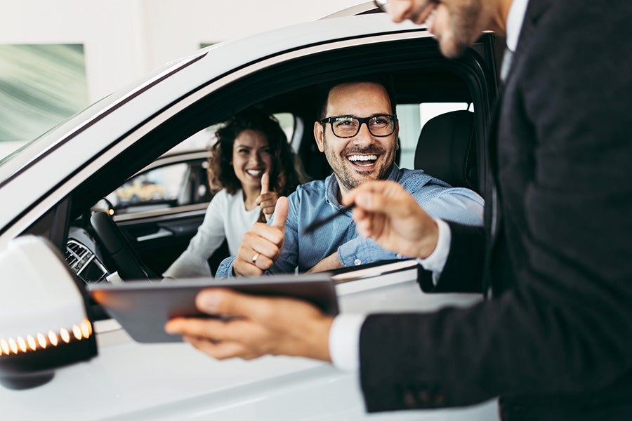 Couple laughing in car while salesman shows info on tablet outside.